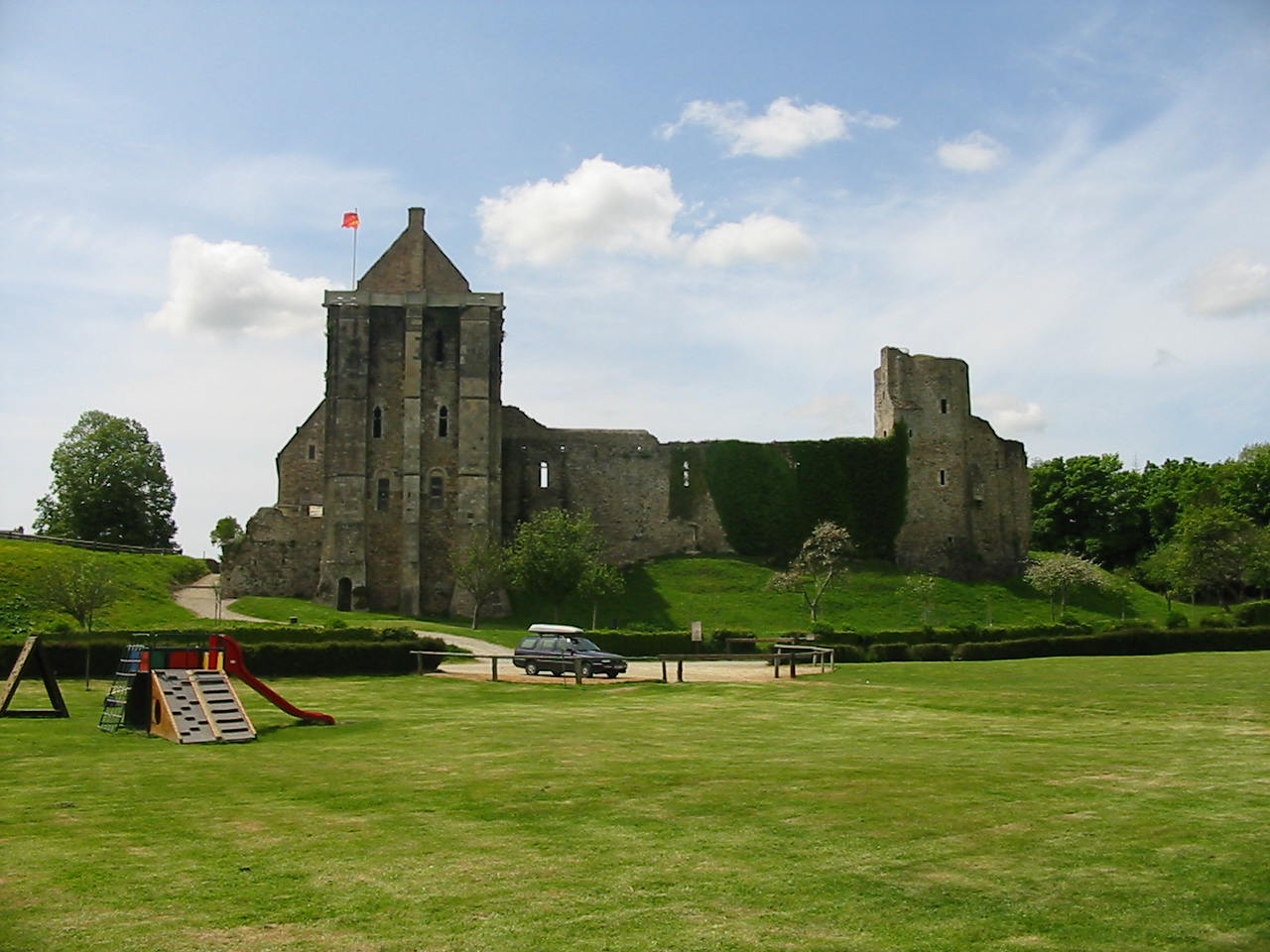 Ruines de château avec tour, voiture, aire de jeux et terrain herbeux.