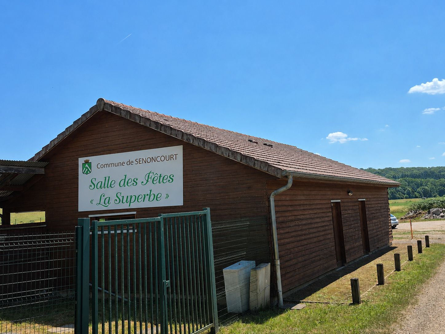 Bâtiment en bois avec une pancarte indiquant "Salle des Fêtes La Superbe" et un portail vert.