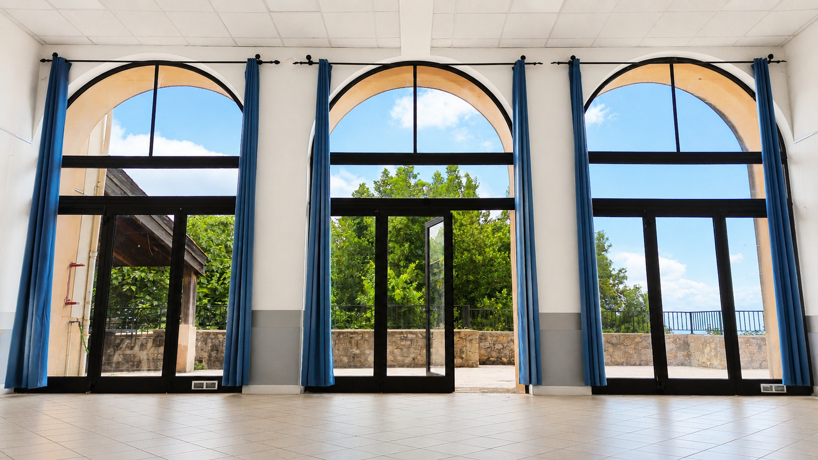Pièce avec trois grandes portes vitrées en arc, rideaux bleus, sol carrelé, vue sur l'extérieur.