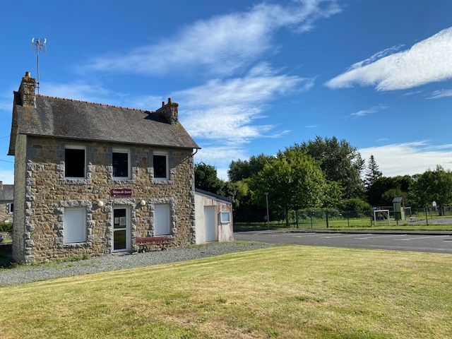 Maison en pierre avec toit en ardoise, antenne sur le toit, banc rouge devant, pelouse et route.