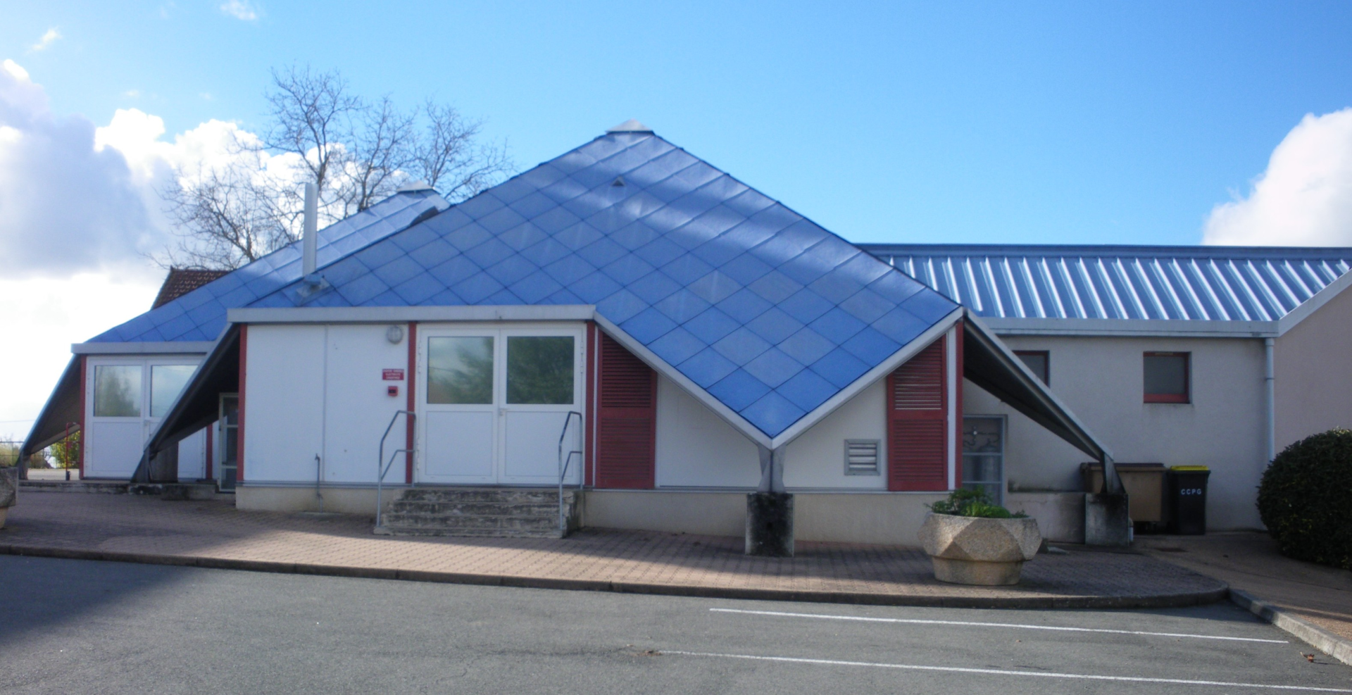 Bâtiment blanc avec toits en acier bleu, portes rouges, fenêtres, escaliers, poubelles et pot de fleur.
