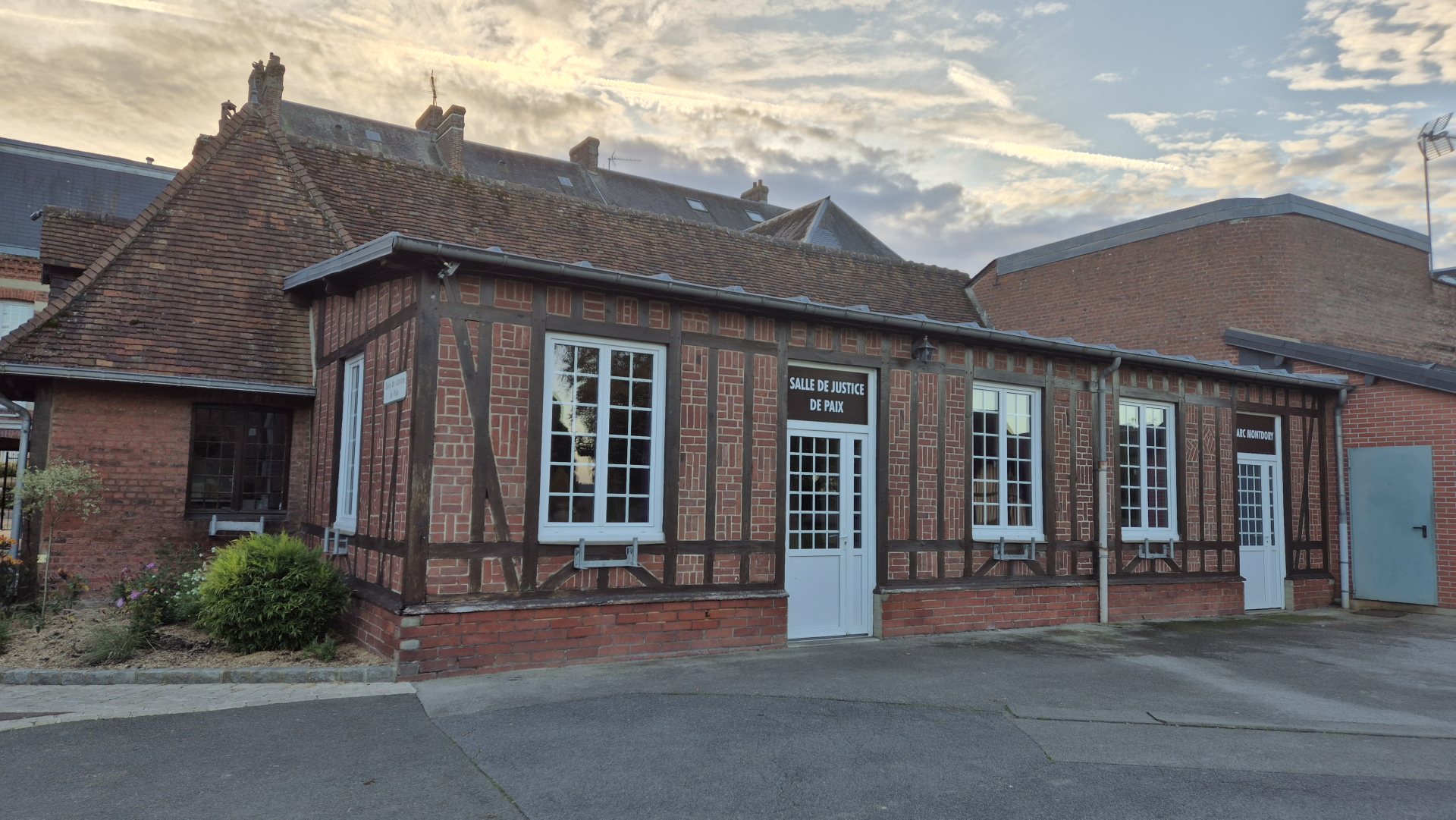 Bâtiment en briques avec toit en tuiles, fenêtres et portes blanches, "Salle de justice de paix" inscrite sur la porte. Cour asphaltée.
