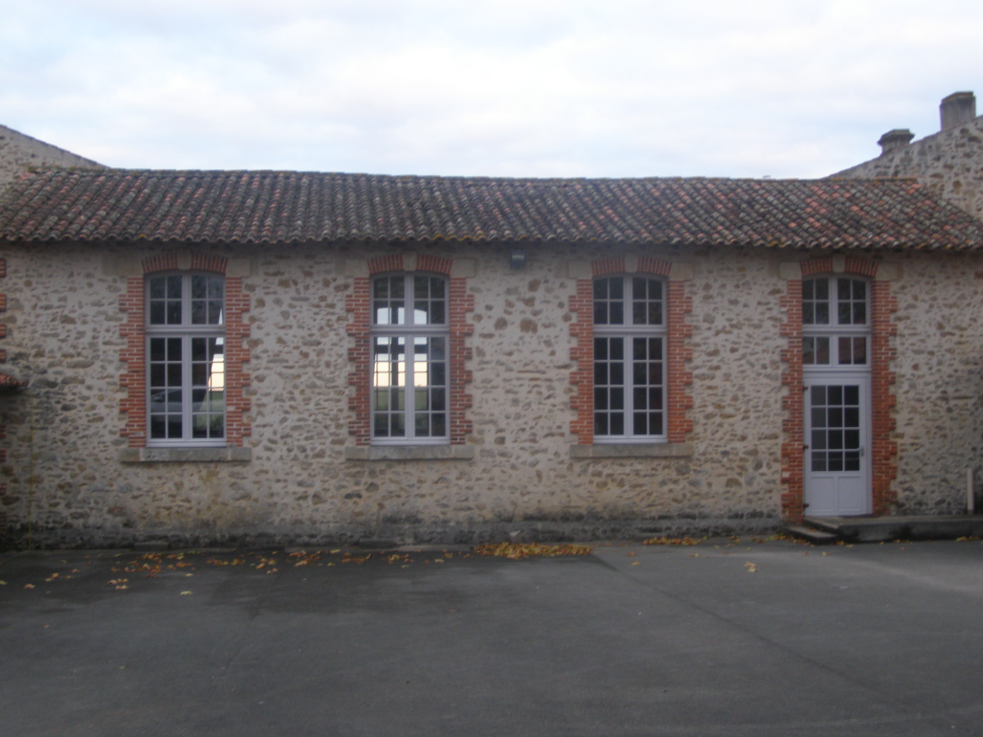 Bâtiment en pierre avec toiture en tuiles, quatre fenêtres et une porte blanche. Cour asphalte.