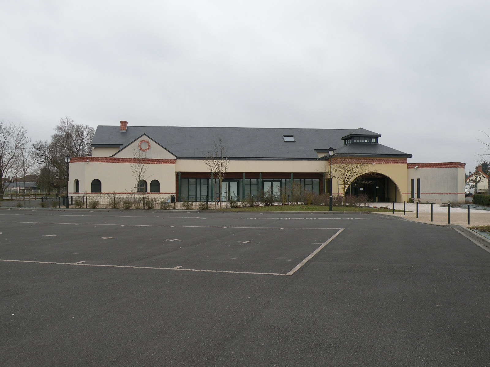 Bâtiment avec façade beige, toiture grise, fenêtres et porte d'entrée. Parking vide devant.
