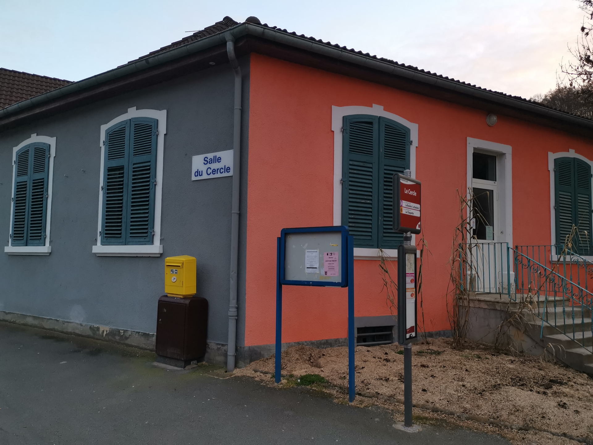 Bâtiment avec façade rose, volets verts, panneau "Salle du Cercle", boîte aux lettres jaune, panneau d'affichage.