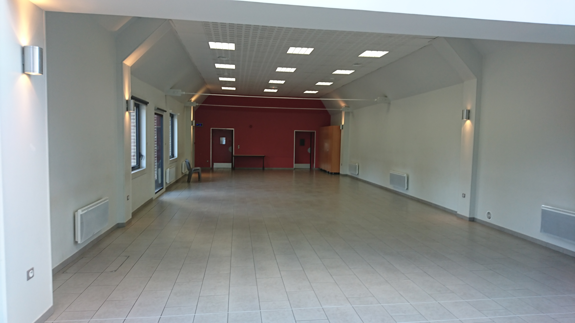Pièce avec plafond haut, murs blancs, sol carrelé, portes, fenêtres, radiateurs et éclairages muraux.