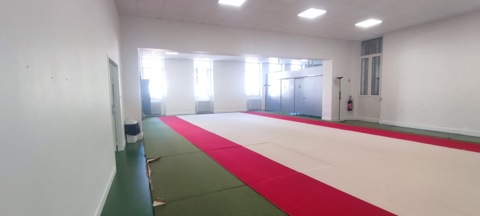 L'image montre une salle de sport vide avec des tapis de sol rouges. Les murs sont blancs, avec des fenêtres et une porte à l'arrière-plan.
