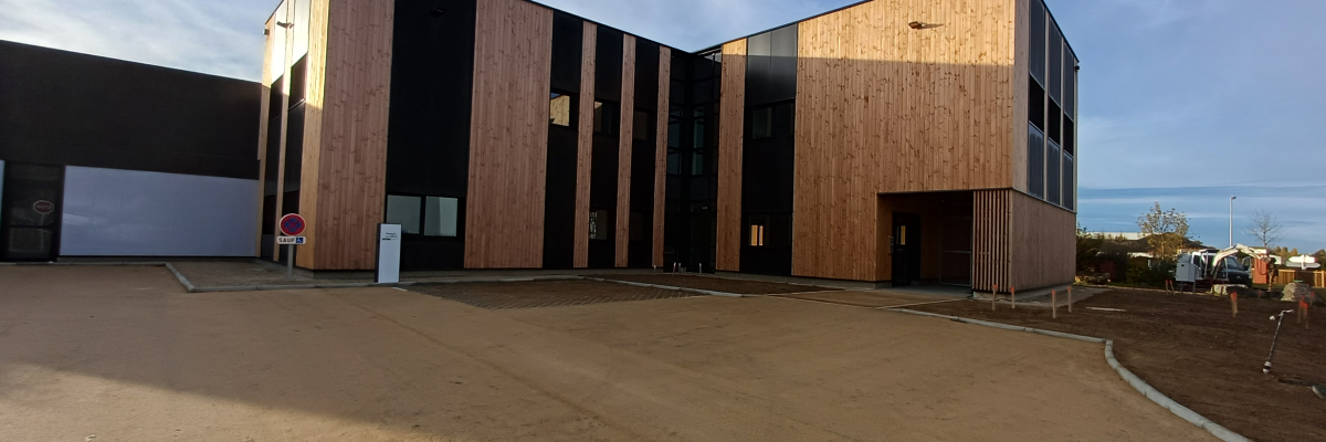 Bâtiment en bois avec grandes fenêtres, parking sableux, ciel bleu.