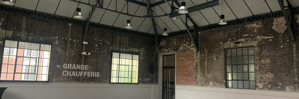 Grande salle au plafond haut avec fenêtres, portes et projecteurs. Sol lisse et murs en brique.