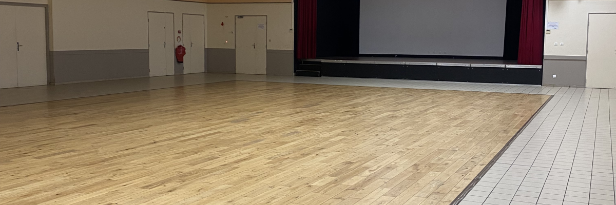 Salle avec plancher en bois, scène, projecteurs, murs blancs et carrelage.