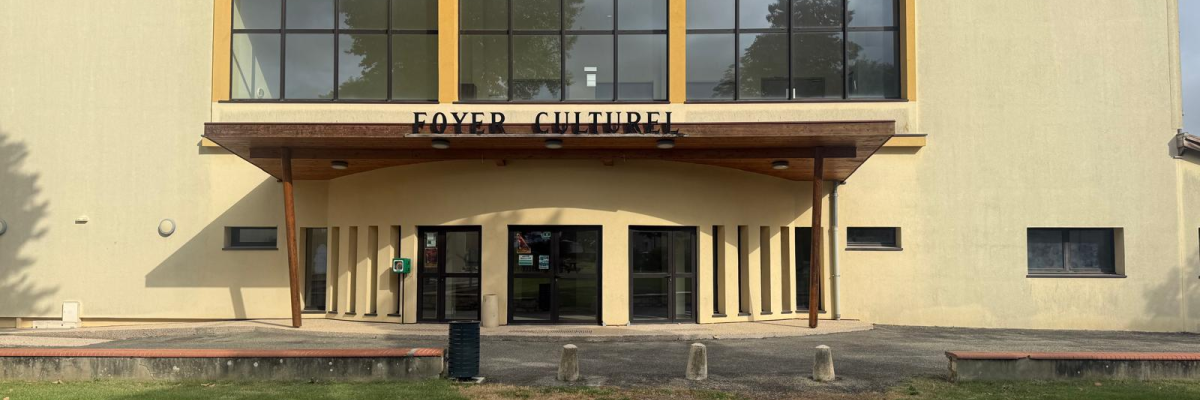 Bâtiment avec entrée, grandes fenêtres, enseigne "Foyer Culturel", pelouse et ciel.