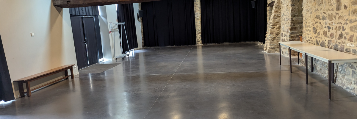 Salle avec tables, bancs, rideaux, sol poli, poutres et pierres apparentes.