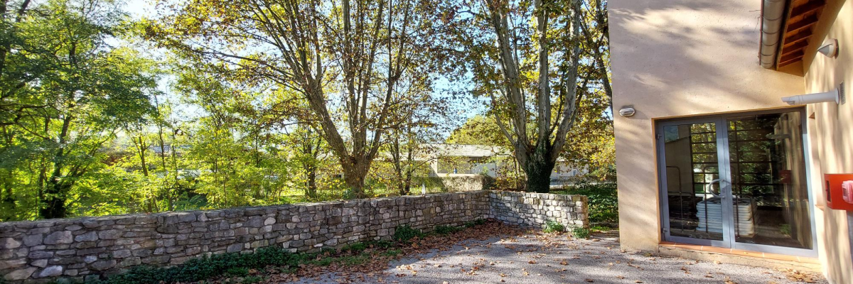 Maison en béton avec entrée en verre, allée en gravier, mur en pierre et arbres feuillus.
