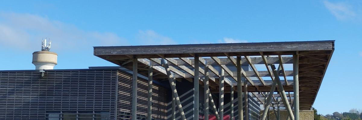 Bâtiment moderne avec structure en bois et façade en verre. Toit en surplomb soutenu par des poteaux métalliques. Ciel bleu en arrière-plan.