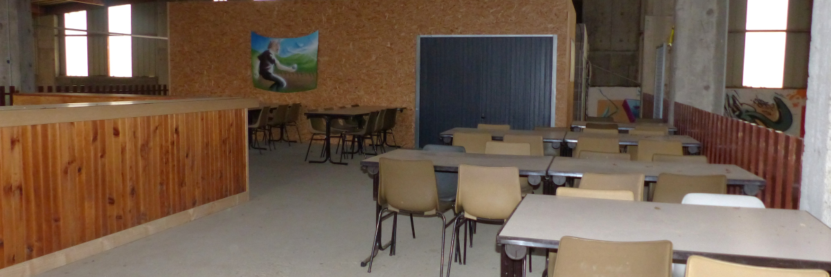 Salle avec tables, chaises, bar en bois, mur en OSB, porte métallique, fenêtres.