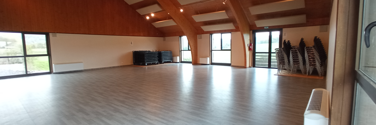 Salle spacieuse avec plancher en bois, grandes fenêtres, chaises empilées, ventilateur, et éclairage encastré.