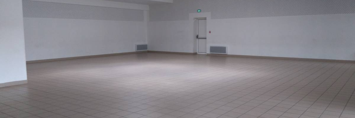 Grande salle vide avec sol carrelé, murs blancs, éclairage au plafond, ventilation et porte.