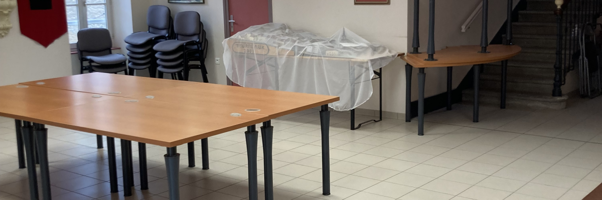 Salle avec tables, chaises, buffet recouvert de nappes, escalier et bannières murales.