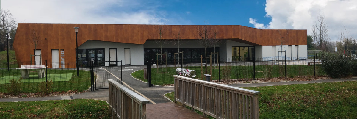 Bâtiment moderne avec façade en métal, entrée principale, rampes d'accès, poteaux et clôture. Vue extérieure, ciel dégagé.