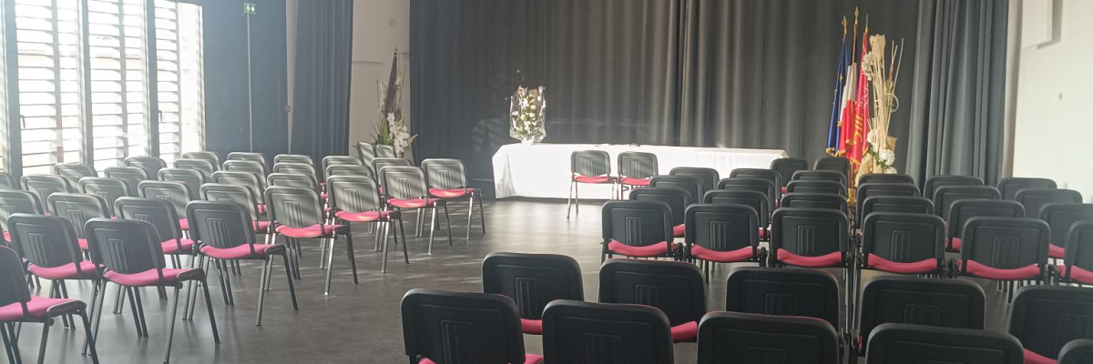 Salle avec chaises alignées, podium, drapeaux, fenêtres et stores.