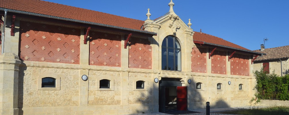 Bâtiment avec toiture en tuiles rouges, murs en pierre et briques, porte centrale rouge et fenêtres de chaque côté.
