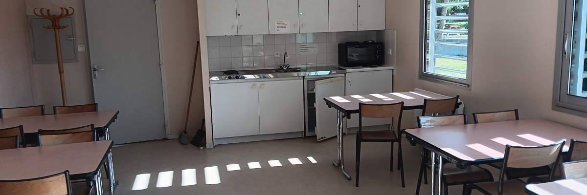 Salle avec tables, chaises, cuisine, évier, micro-ondes, placards et porte-manteau.