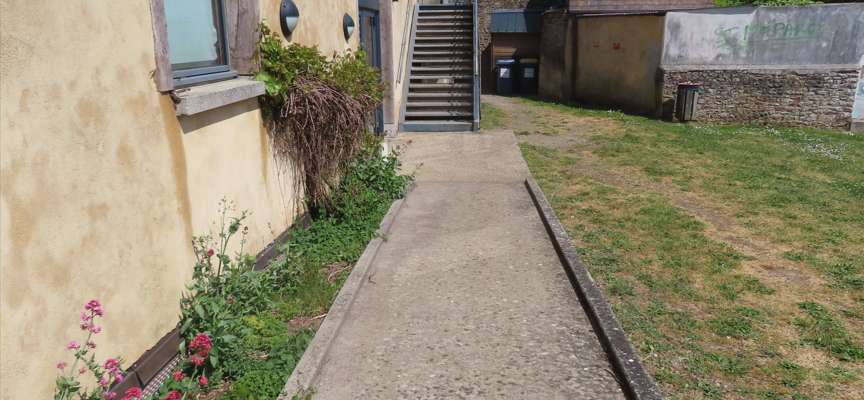 Allée en béton menant à un escalier extérieur, bordée de fleurs et d'herbes, avec des bâtiments en pierre et en brique de chaque côté.