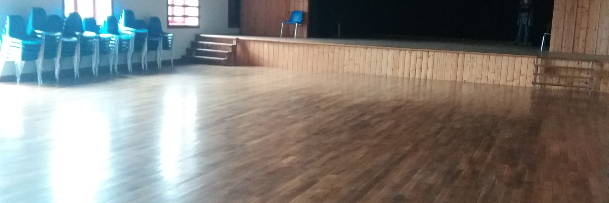 Salle vide avec parquet, chaises bleues, scène, rideau noir, lumières suspendues et murs en béton.