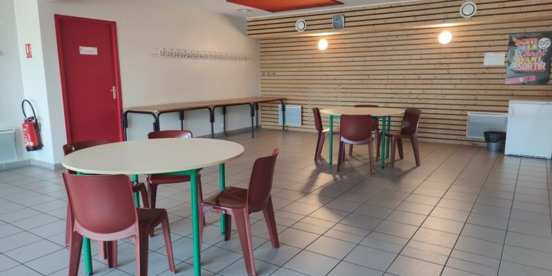 Salle avec tables rondes, chaises, porte rouge, extinction et affiche sur mur lambrissé.