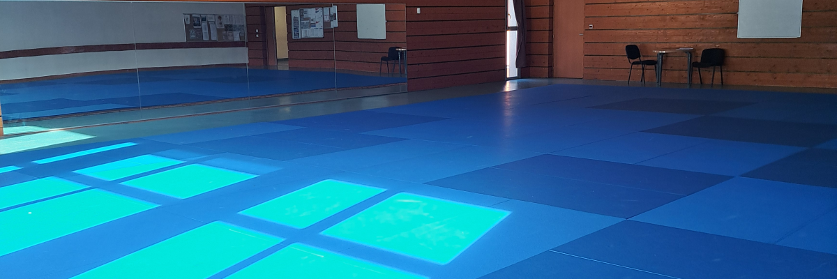 Salle polyvalente avec sol bleu, murs en bois, tables, chaises et projecteurs suspendus.