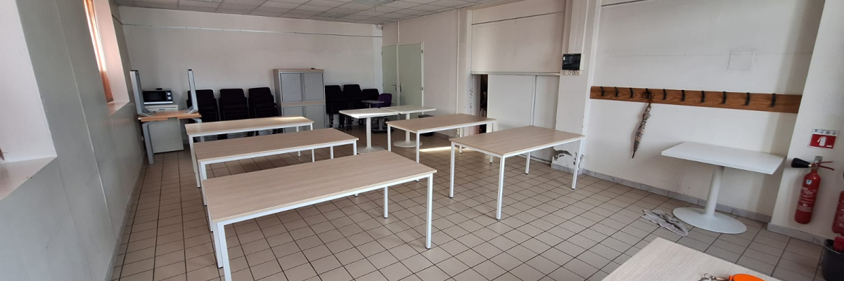 Salle de classe avec tables, chaises, tableau blanc et extincteur.