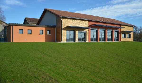 Bâtiment rectangulaire avec toiture en tuiles rouges. Façade en briques avec sections orange et blanches. Grandes fenêtres et portes vitrées. Pelouse verte devant.