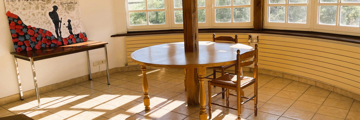 Salle avec table ronde, chaises, banc, tableau, fenêtres et poutre en bois.