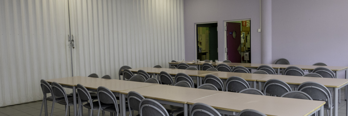 Salle de classe avec tables, chaises, tableau blanc, porte et éclairage.