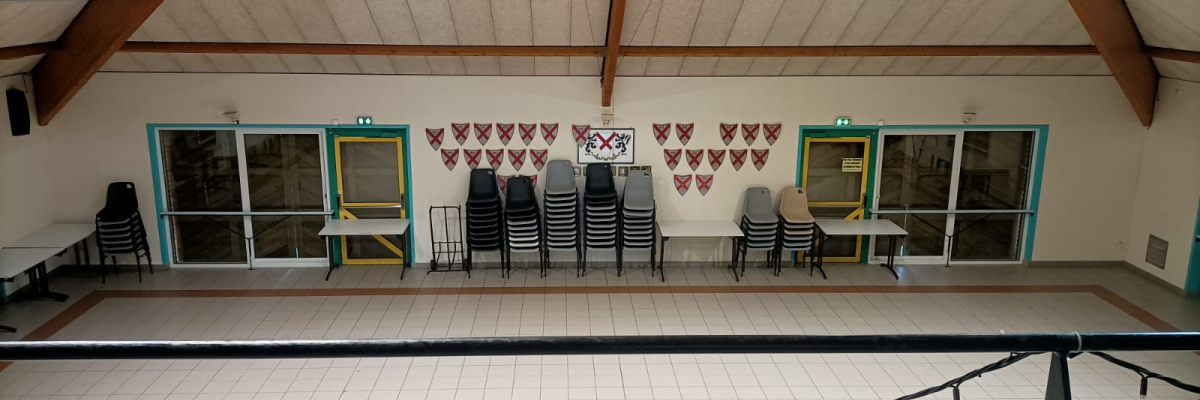Salle polyvalente avec chaises empilées, tables pliantes, grandes fenêtres et décor mural.