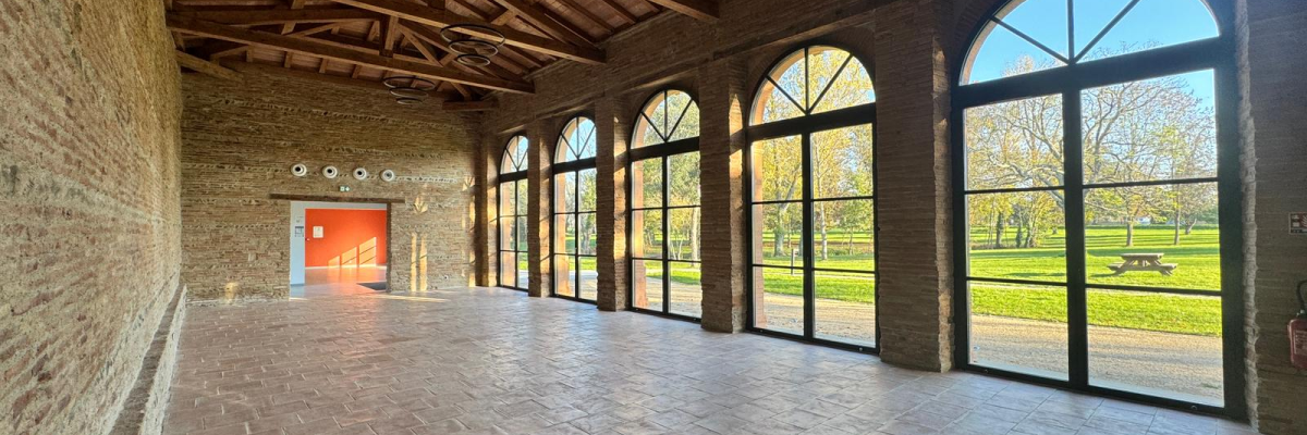Salle avec murs en brique, sol en carreaux, grandes fenêtres, porte rouge et plafond en bois.