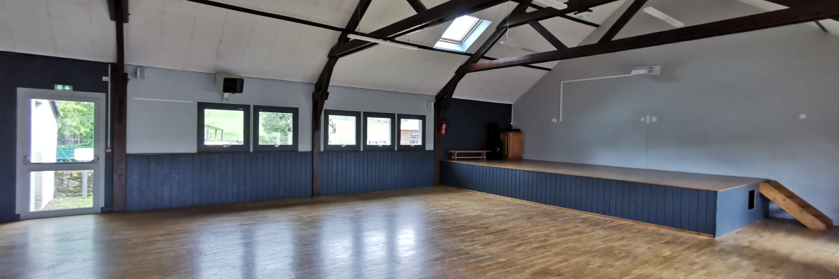 Salle avec plancher en bois, murs bleus, fenêtres, porte, escalier et poutres apparentes.