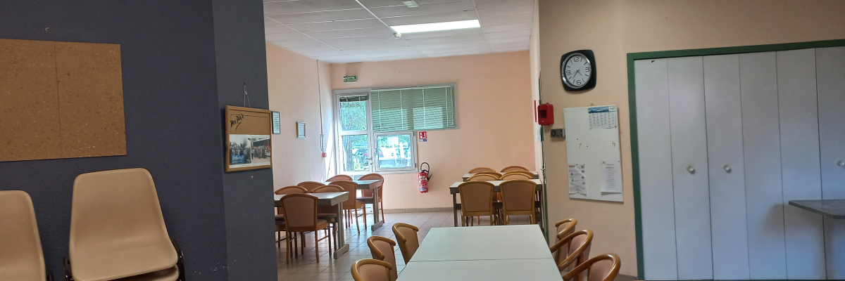 Salle avec tables, chaises, tableau, horloge, extinguisseur, armoire et fenêtre.