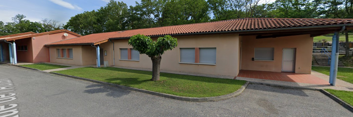 Bâtiment avec toiture en tuiles, murs crème et portes bleues. Arbre et pelouse devant, route et parking à proximité.