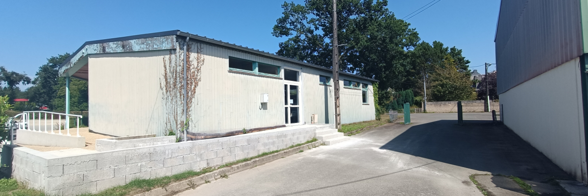 Bâtiment bas avec façade blanche, toit gris, rampes d'accès, portes vitrées, arbres et route asphaltée.