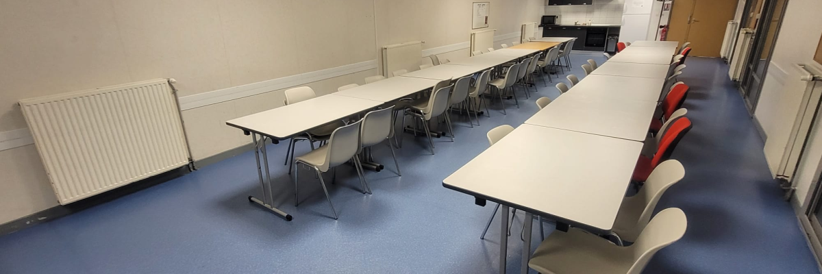 Salle avec tables, chaises, radiateur, télévision, cuisine et porte.