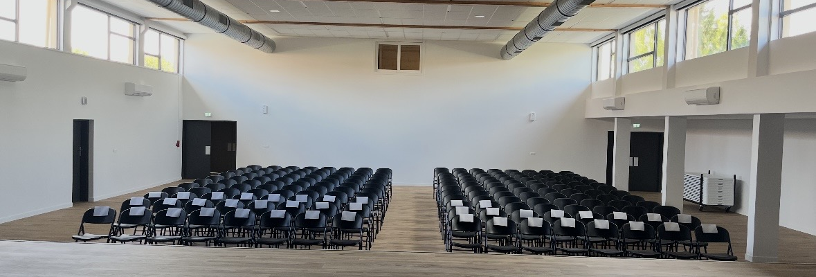 Salle avec rangées de chaises, sol en bois, murs blancs, fenêtres, portes et conduits au plafond.