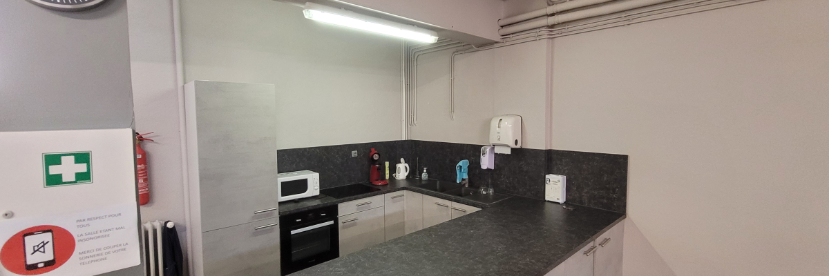 Cuisine avec plan de travail, étagères, micro-ondes, four, panneaux électriques, extincteur, affiches de sécurité, horloge murale.