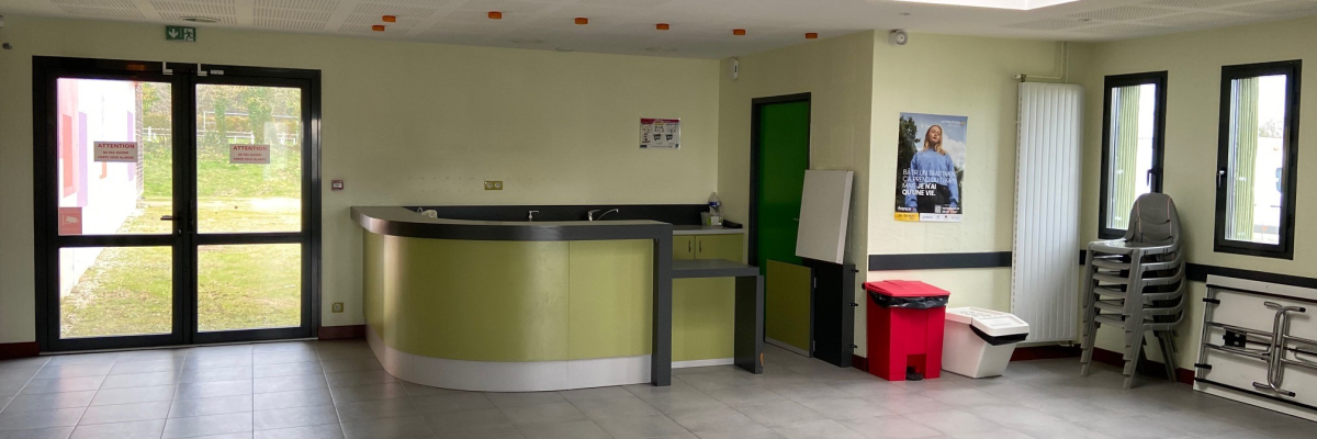 Réception avec comptoir, portes, affiches, poubelles, chariot, escalier et sol carrelé.