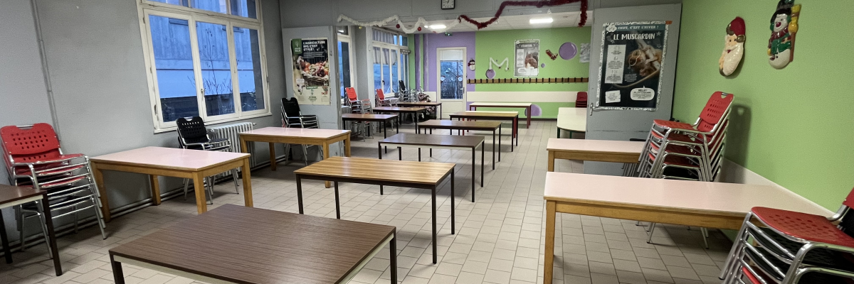 Salle de classe avec tables, chaises, tableau blanc, étagères, fenêtres et mobilier empilé.
