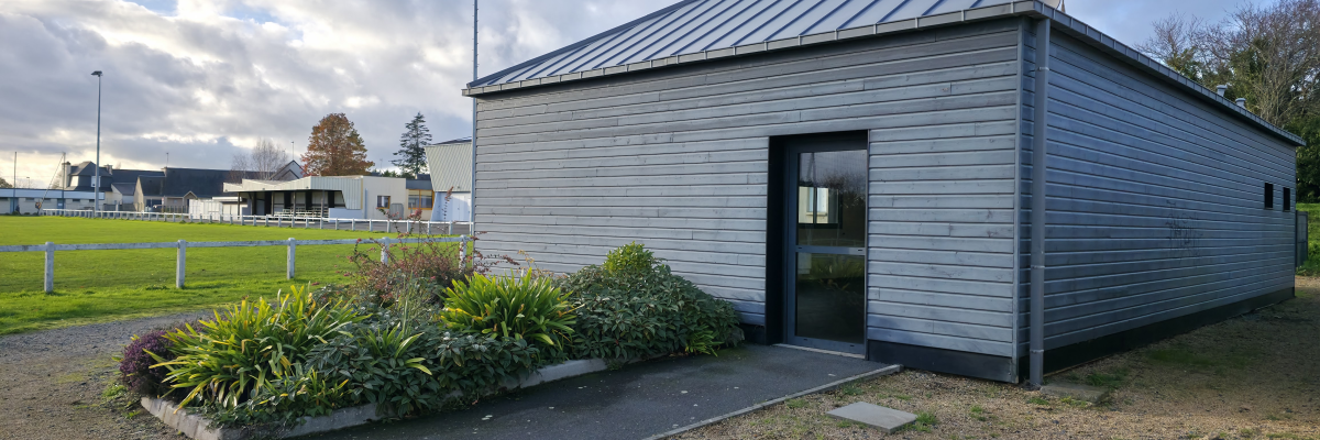 Petit bâtiment rectangulaire en bois gris avec toit plat et porte au milieu. Jardinières avec des plantes vertes devant l'entrée. Ciel partiellement nuageux en arrière-plan.