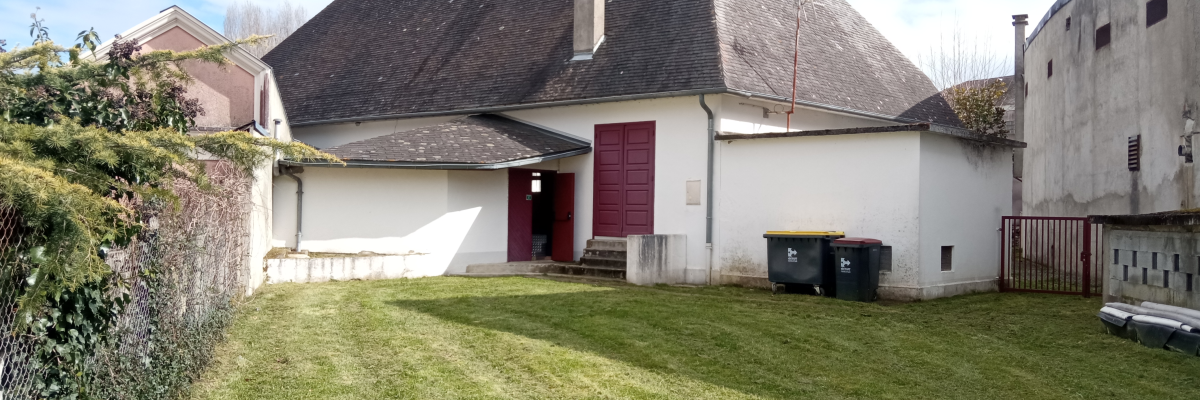 Maison blanche avec toit en ardoise, porte rouge, poubelles, pelouse, haie et mur en brique.