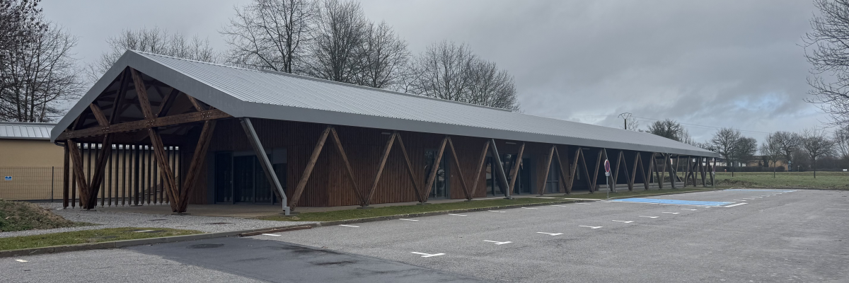 Bâtiment long avec toit incliné, structure en bois et grandes fenêtres, stationnement devant.