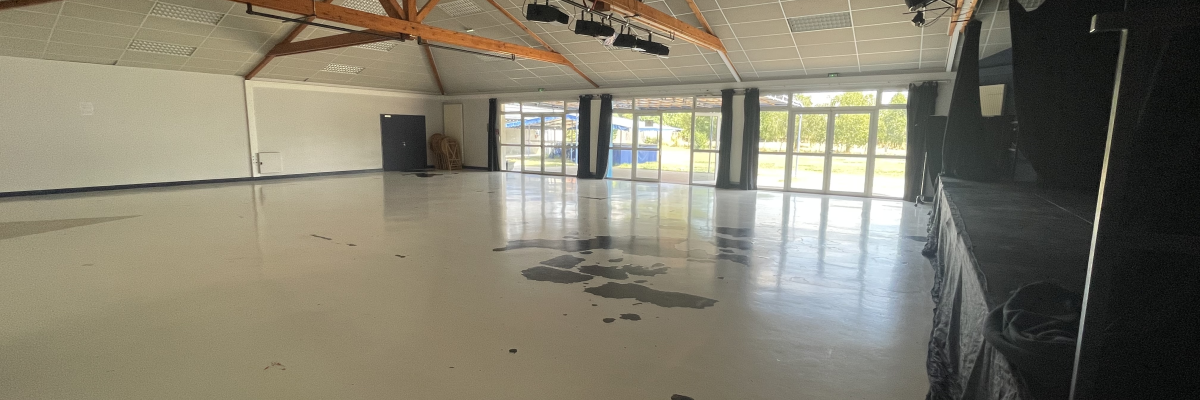 Salle vide avec grandes fenêtres, poutrelles apparentes, spots et ventilateur au plafond.