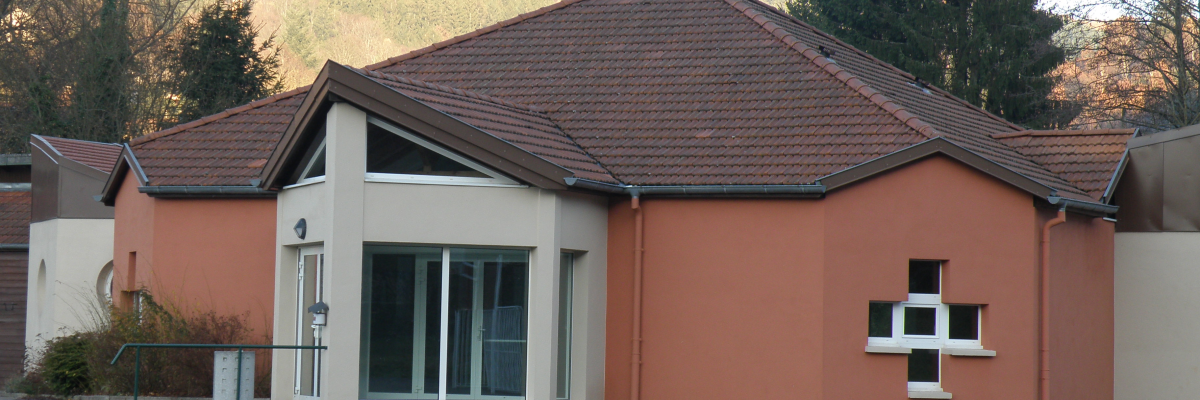 Bâtiment avec toit en pente, murs peints en rose et blanc, fenêtres, porte vitrée, et une croix en brique sur le mur.